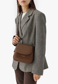 Pruuni nahast õlavarre kott tekstureeritud klapiga, hõbedase logo ja kitsega rihmaga, mida hoiab inimene hallis bleiseris.