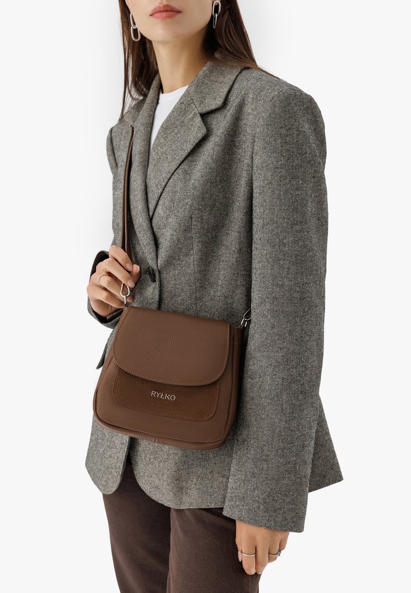 Pruuni nahast õlavarre kott tekstureeritud klapiga, hõbedase logo ja kitsega rihmaga, mida hoiab inimene hallis bleiseris.