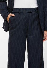 Pantalon de costume bleu marine à rayures fines avec poches latérales, porté avec un blazer assorti et un haut blanc, main reposant dans la poche gauche.