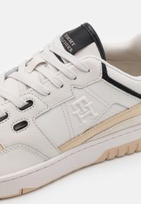 Sneaker in pelle bianca con dettagli neri, striscia beige e suola testurizzata. Presenta cuciture visibili e logo sulla linguetta. Design classico con lacci.