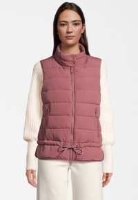 Padded vest w jasnym mauve z wysokim kołnierzem, zamkiem z przodu i regulowanym ściągaczem w pasie. W zestawieniu z kremowym swetrem.