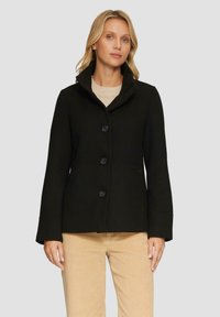 Schwarze Wollmischungjacke mit Kragen, vier Knöpfen und Seitentaschen, kombiniert mit einem hellen Pullover und beigen Cordhosen.