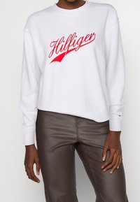 Sudadera blanca con el logo "Hilfiger" en rojo. Presenta un cuello redondo, puños acanalados y un corte relajado. Se lleva con pantalones de cuero sintético marrones.