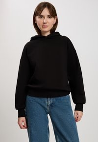 Czarna bluzka z kapturem o luźnym kroju, z prążkowanymi mankietami i teksturowanym logo na środku. Połączona z jasnoniebieskimi, wyblakłymi dżinsami.