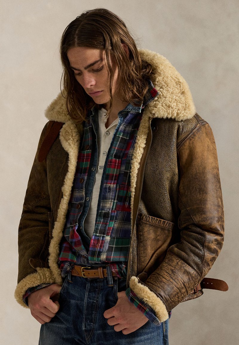 Giovane uomo con capelli lunghi che indossa una giacca di shearling marrone sopra una camicia a quadri e jeans, guarda verso il basso con le mani vicino alle tasche.
