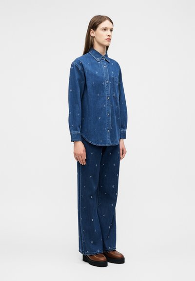 Une femme se tient de profil droit, portant une chemise en jean bleue et un pantalon large orné de petits motifs blancs, assorti à des chaussures compensées marron.