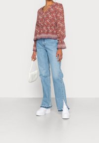 Blusa rossa floreale con maniche a campana, abbinata a jeans dritti azzurri chiari con spacchi laterali, e completata da una borsa bianca.