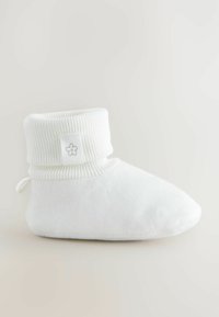 Chausson en peluche blanc avec un revers côtelé, présentant un petit emblème de fleur sur le côté et un design de bout arrondi et doux.