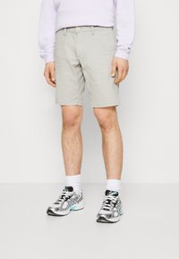 Shorts gris clair avec un design à l'avant plat, fermeture à bouton et poches latérales. Portés avec des chaussettes blanches et des chaussures de sport noir et argent.