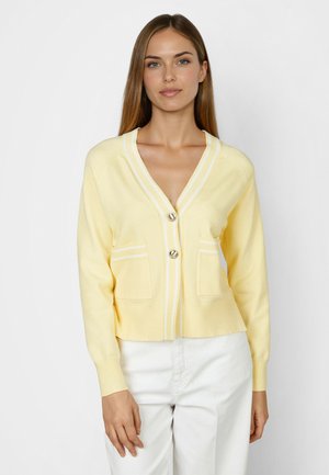 Woman wearing a light yellow buttoned cardigan with white stripes on pockets and cuffs, paired with white pants, standing against a plain background.