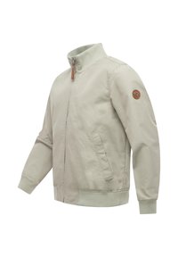 Beige kevyt takki resorikauluksella, -hihojen suulla ja helmassa, vetoketjulla edessä, sivutaskuilla ja pyöreällä nahkaisella logopatchilla vasemmassa hihassa.