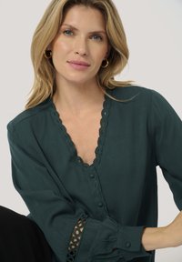 Blusa verde acqua con scollatura a scallop e dettagli con bottoni, con accenti in pizzo sulle maniche. Realizzata in tessuto leggero con una vestibilità morbida.