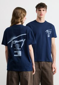 Δύο μοντέλα φορούν μπλουζάκια Tommy Jeans σε ναυτικό μπλε, το ένα παρουσιάζει το λογότυπο μπροστά και το άλλο το μεγάλο λογότυπο στην πλάτη με ονόματα πόλεων.