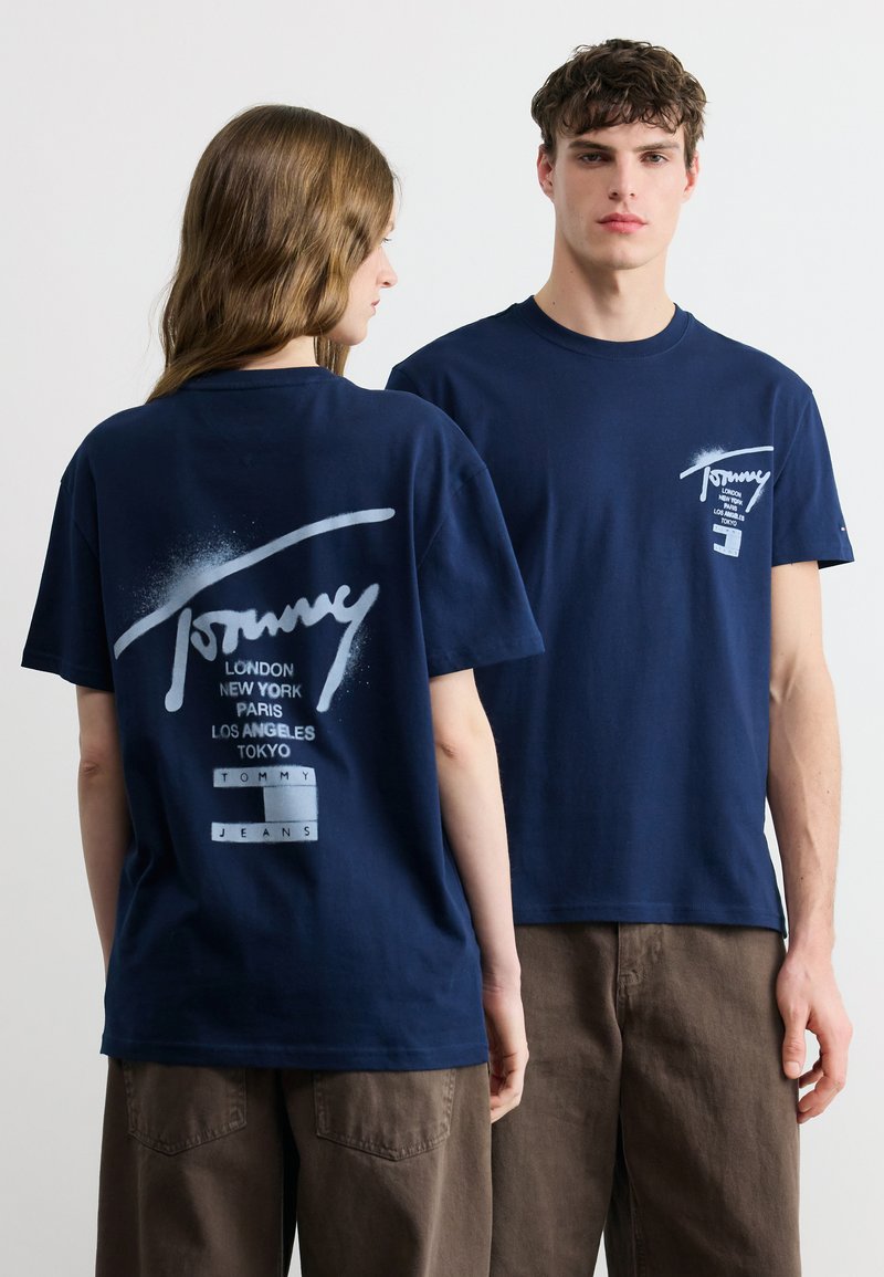 Δύο μοντέλα φορούν μπλουζάκια Tommy Jeans σε ναυτικό μπλε, το ένα παρουσιάζει το λογότυπο μπροστά και το άλλο το μεγάλο λογότυπο στην πλάτη με ονόματα πόλεων.