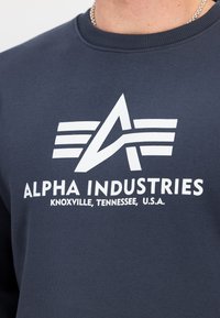 Jūras spēku kokvilnas džemperis ar lielu baltu "Alpha Industries" logo un stilizētu "A" un horizontālām svītrām. Ar apaļu apkakli.