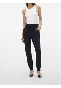 Pantalons noirs taille haute avec une ceinture nouée, présentant une texture lisse et des poches latérales. Associés à un débardeur blanc ajusté et des chaussures noires.