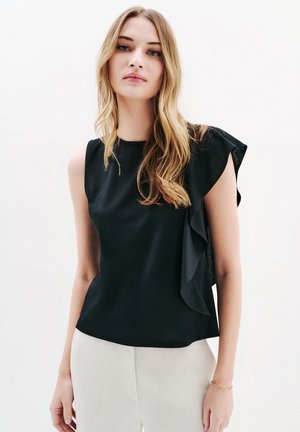 Zwarte mouwloze blouse met een watervalruche op de schouder; gladde stof met een getailleerde pasvorm, gecombineerd met lichte broek.