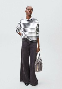 Nainen, jolla on päällä raitapaitapusero ja leveälahkeiset mustat farkut, pitää harmaasta käsilaukusta, jossa on kultainen avainrenkaat, seisten yksinkertaista taustaa vasten.