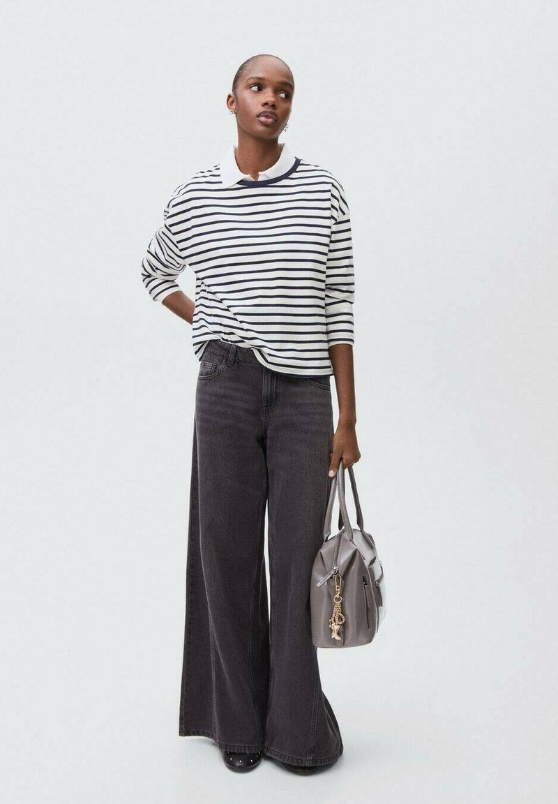 Nainen, jolla on päällä raitapaitapusero ja leveälahkeiset mustat farkut, pitää harmaasta käsilaukusta, jossa on kultainen avainrenkaat, seisten yksinkertaista taustaa vasten.