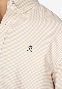 Camisa de cuadros beige con collar, que presenta un pequeño emblema en color azul marino. El material tiene un patrón de cuadrícula texturizado; los botones son de color claro.