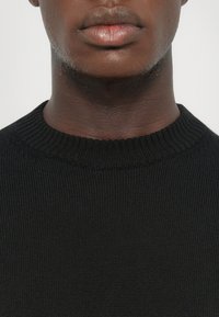 Schwarzer Strickpullover mit geripptem Rundhalsausschnitt und strukturiertem Stoff, der die Nahaufnahme des Halses und der Lippen hervorhebt.