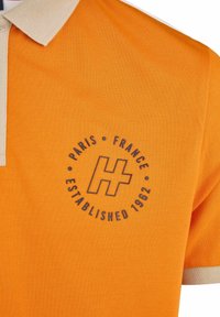 Polo orange avec un col beige, un logo circulaire indiquant "Paris France Établi en 1962" et un tissu texturé.