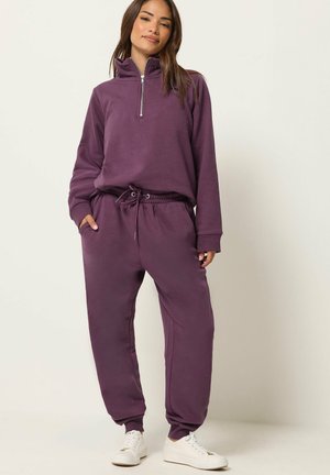 Kvinde iført en lilla sweatshirt med halv lynlås og matchende joggebukser, stående med hænderne i lommerne, hvide sneakers.