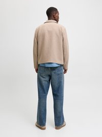 Rjav jekleni jopič iz volnenega materiala s ovratnikom, v kombinaciji z modrimi kavbojkami. Obleka je dopolnjena s svetlimi čevlji, prikazana s hrbtne strani.