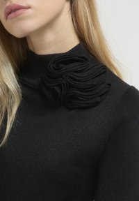 Donna che indossa un top nero a collo alto con un grande dettaglio floreale in tessuto nero sulla spalla, capelli biondi visibili contro uno sfondo chiaro.