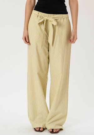 Pantaloni a gamba larga a righe verticali gialle e bianche con cintura annodata in vita, indossati con top nero e sandali marroni con punta aperta.