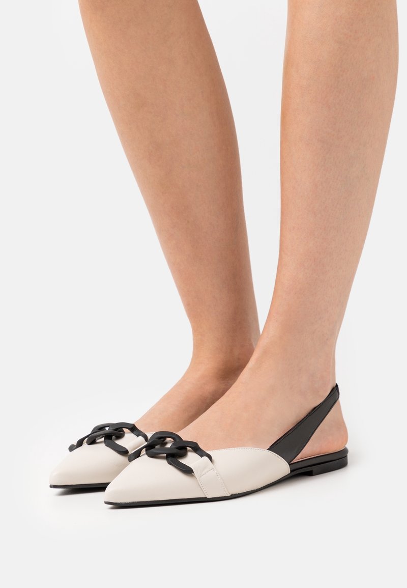 Platta slingback-skor i vit och svart läder, med spetsig tå, kedjeaccent framtill och en minimalistisk design.