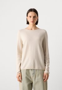 Beige langermet topp i myk strikket stoff med rund hals. Har minimale sømdetaljer og en avslappet passform.