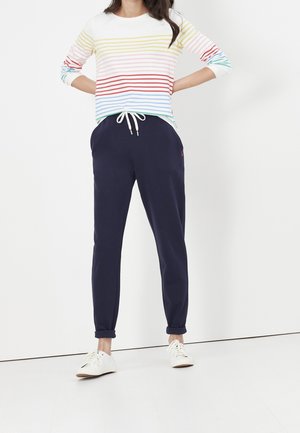 Femme portant une chemise blanche à manches longues avec des rayures multicolores et un pantalon bleu marine à cordon avec des baskets blanches sur fond blanc.