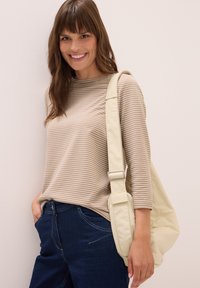 Lächelnde Frau in beigem und weiß gestreiftem Oberteil, dunkblauen Jeans, die eine krämfarbene Schultertasche vor einer schlichten Wand trägt.