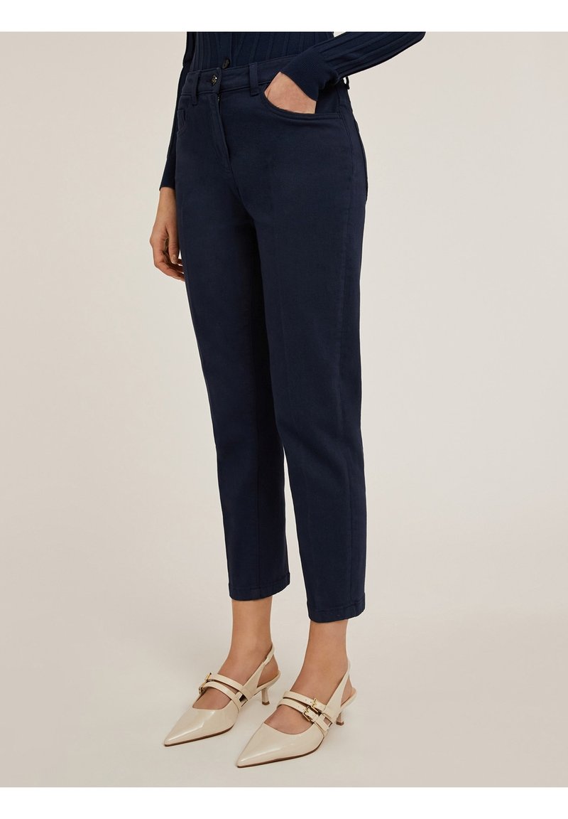 Pantalon bleu marine fuselé avec poches avant, fermeture par bouton, et une texture lisse, associé à des chaussures slingback beiges.