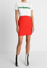 Bílý tričko s zeleným textem, spárované s červenou strukturovanou mini sukní. Černé a bílé boty na vysokém podpatku dokončují outfit.