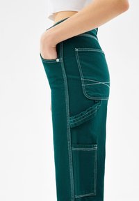 Nainen, jolla on yllään korkeavyötäröiset tummanvihreät cargo-housut valkoisilla ompeleilla, käsi etutaskussa ja näkyvät sivutaskut, valkoista taustaa vasten.