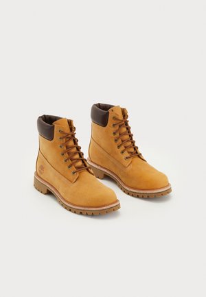 Par de botas de trabalho de pele bege com colarinhos acolchoados castanhos, ilhós de metal e solas robustas de ranhura sobre um fundo branco.