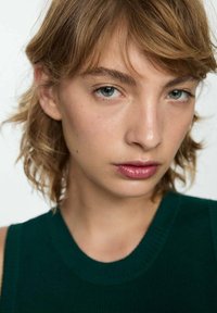 Primo piano di una persona con capelli castano chiaro e pelle chiara, che indossa un top verde senza maniche. Le caratteristiche includono occhi blu chiari e pelle liscia.