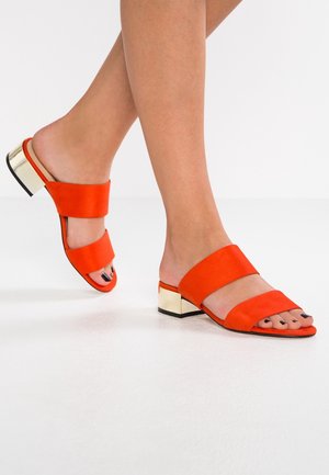 Jambes portant des sandales à talon bloc bas, à double bride, en daim orange à bout ouvert avec talons métalliques dorés sur un sol blanc.