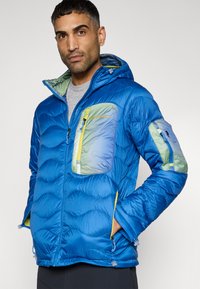 Μπλε μπουφάν τύπου puffer με κουκούλα, διακοσμημένο με κυματιστό σχέδιο, τσέπες με πράσινες και μπλε λεπτομέρειες, φερμουάρ και ελαστικές μανσέτες. Ελαφρύ και μονωμένο.