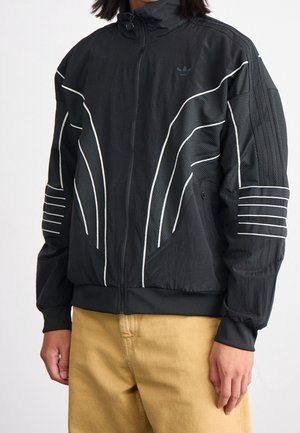 Personne portant une veste noire zippée avec des lignes blanches incurvées et des rayures sur les manches, associée à un pantalon marron clair, sur fond blanc.