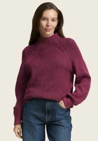 Strickpullover inweinrot mit gerippter Textur und hohem Kragen, mit Raglanärmeln. Kombiniert mit dunkelblauen Jeans. Einfacher, lässiger Stil.