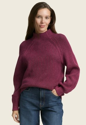 Bordeaux gebreid vest met ribbelstructuur en hoge kraag, voorzien van raglanmouwen. Gepaarde met donkerblauwe jeans. Eenvoudige, casual stijl.