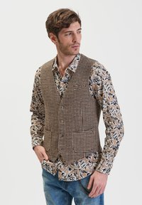 Gilet in tessuto a motivo houndstooth marrone con cinque bottoni, indossato sopra una camicia a fantasia floreale beige e nera, abbinato a jeans blu.