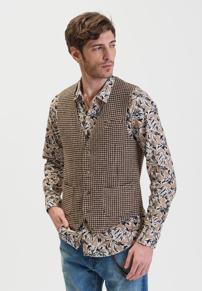 Gilet in tessuto a motivo houndstooth marrone con cinque bottoni, indossato sopra una camicia a fantasia floreale beige e nera, abbinato a jeans blu.