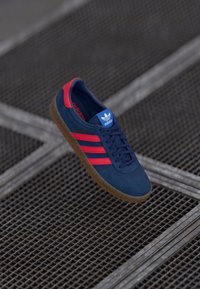 Blå ruskinds sneakers med røde detaljer, der har tre striber, en tekstureret gummisål og en blå tunge med et logo.