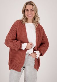 Gebreide cardigan in roestkleur met een open voorkant, losse pasvorm en textuurpatroon. Gecombineerd met een witte top en gestreepte broek.