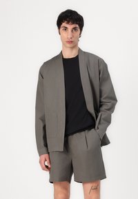 Grå bomulljakke med én lomme, sammen med matchende shorts. Modellen har på seg en svart skjorte under. Minimalistisk design, avslappet passform.