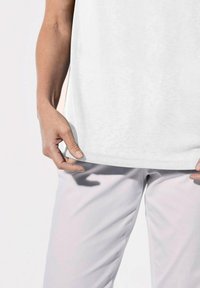 Camicia bianca a maniche corte realizzata in tessuto morbido, con una texture liscia. Una mano tiene l'orlo della camicia, indossata con pantaloni chiari.